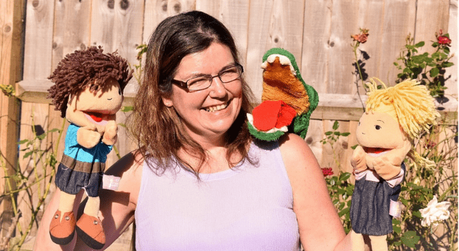 Cath stands in a sunny garden holding her Sam and Sally puppets, with Mr Crocodile puppet sitting on her shoulder.