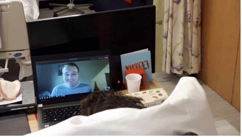 A patient in a hospital bed looks at a laptop screen showing the face of the student SLT in one of their virtual sessions